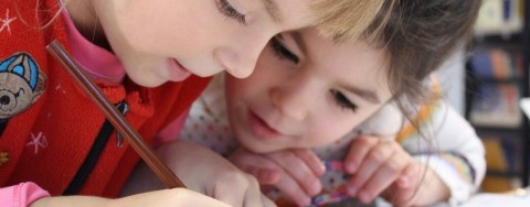Auf dem Bild sind zwei Kinder zu sehen, ein blonder Junge und ein braunhaariges Mädchen. Der Junge trägt einen roten Pullover auf dessen rechter Seite ein Bär abgebildet ist. Er hält einen dunkelroten Stift in der rechten Hand. Er scheint etwas zu schreiben oder zu zeichnen. Das Mädchen beugt sich zu ihm rüber und schaut gespannt, was er schreibt oder zeichnet.