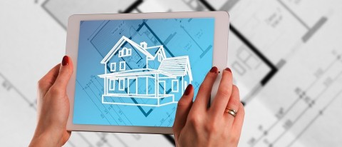 Auf dem Bild sind die Hände einer Frau zu sehen. Sie trägt roten Nagellack und am rechten Ringfinger einen breiten Ring. Sie hält ein weißes Tablet in den Händen, auf dem die Skizze eines Hauses zu sehen ist. Der Bildschirmhintergrund ist blau und zeigt den Grundriss eines Gebäudes. Der Grundriss liegt auch nochmal in Papierform auf einem Tisch unter dem Tablet. Die Skizze des Hauses wirkt wie eine Projektion des fertigen Gebäudes über dem Plan des Gebäudegrundrisses.
