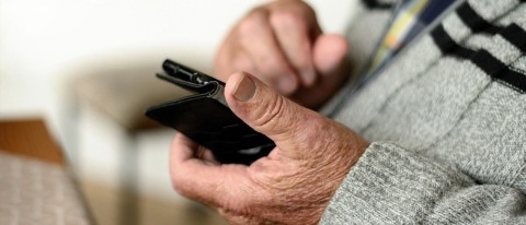 Auf dem Bild sind die Hände eines alten Mannes zu sehen. Er trägt einen grauen Pullover mit zwei schwarzen Streifen über der Brust und hält ein Handy in den Händen. Er scheint darauf zu schauen. Im Hintergrund sind altmodische Möbel zu sehen.