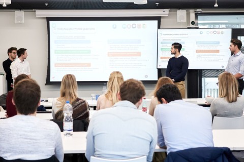 In the photo, four young men in smart clothes can be seen in the background giving a presentation. In the foreground are other people of the same age, also in shirts and blouses, sitting in rows at tables attentively following the presentation. In the photo, four young men in smart clothes can be seen in the background giving a presentation. In the foreground are other people of the same age, also in shirts and blouses, sitting in rows at tables attentively following the presentation.