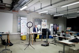 Moderator steht in einem großen Raum an einem Tisch mit großem Bildschirm