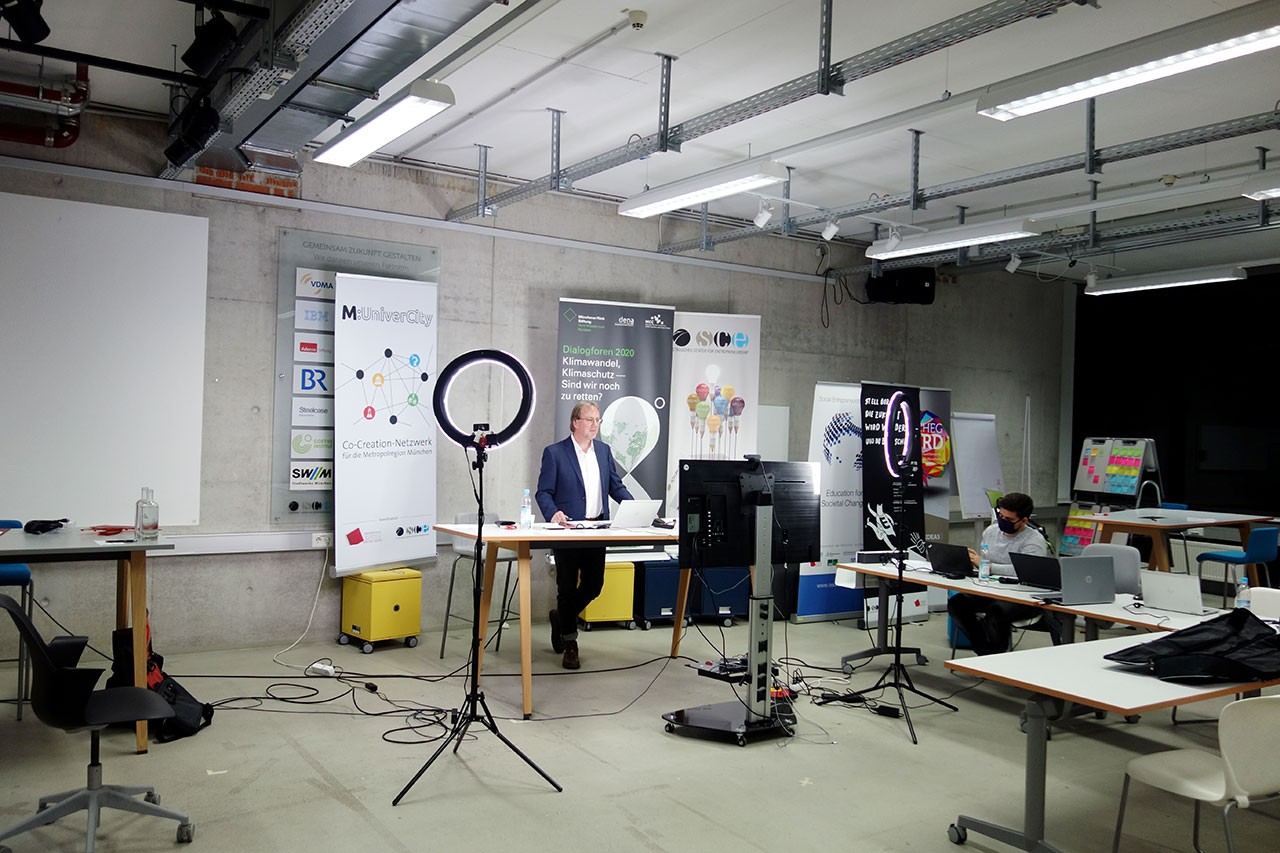 Moderator steht in einem großen Raum an einem Tisch mit großem Bildschirm