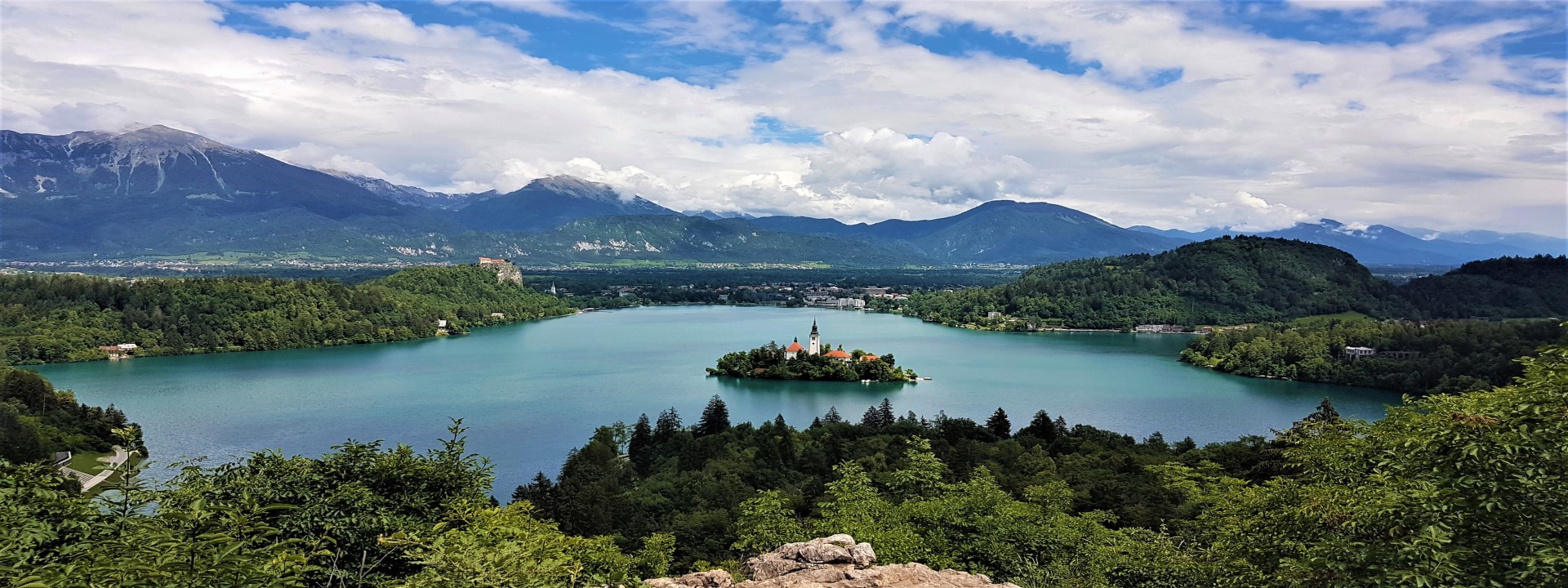 Blick von einem Hügel auf den See in Bled: ein See umrandet mit Bäumen, in der Mitte des Sees eine kleiner Inselt mit einer Kirche. Hinten am See sieht man die Stadt Bled und die Festung. Im Hintergrund Gebirge in Wolken.