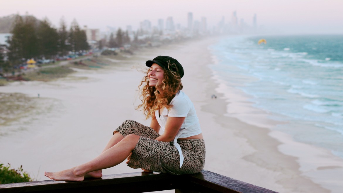 Surfers Paradise in Australien (Sarah Kratzl) Eine lächelnde Studentin mit langen lockingen Haaren sitzt auf einem Balken. Sie hat ein schwarzes Cappy, weißes T-Shirt und Hose mit Leopardenmuster an. Im Hintergrund ein Sandstrand und Ozean sowie eine Großstadt mit Wolkenkratzern.