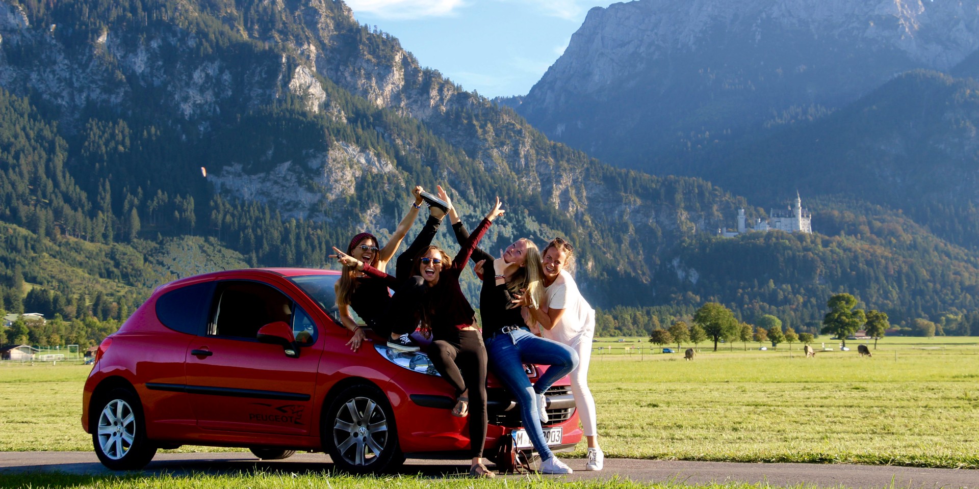 Vier Studentinnen posieren auf der Maske eines roten Kleinwagens, im Hintergrund eine Wiese, Gebirge und Schloss Neuschwanstein