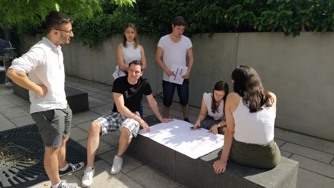 2023_Pers.Fuehrunsgpot_Teamwork (Foto: Hochschule München) Ein Studierenden-Team sitzt mit einem Flipchart-Papier vor dem T-Gebäude.